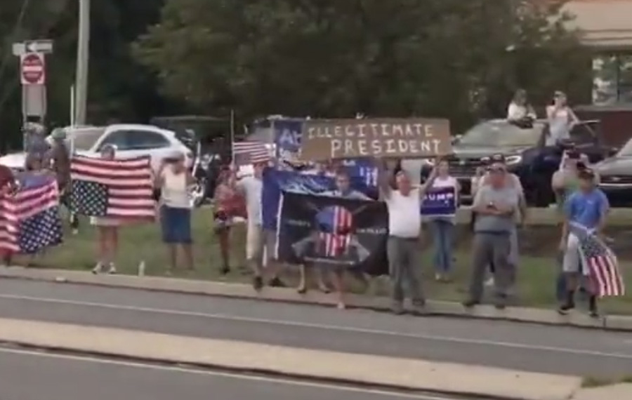 A man holds high a sign saying Illegitimate President as others hold upside down American flags, generally a signal of dire distress in instances of extreme danger to life or property.