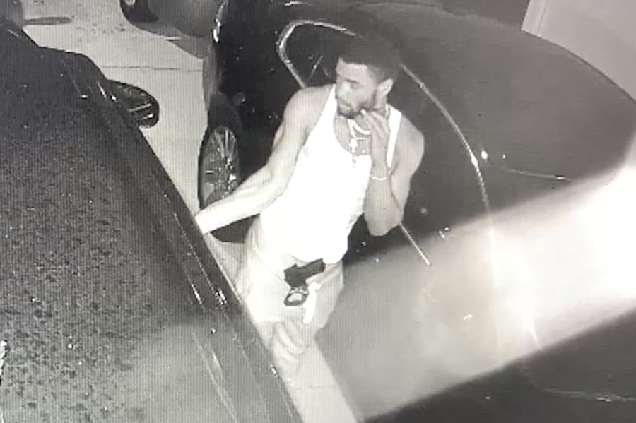  The video shows a young male wearing a white tank top, light colored pants and several necklaces around his neck with a short dark beard, short dark hair and a black handgun in the front of his waistband.